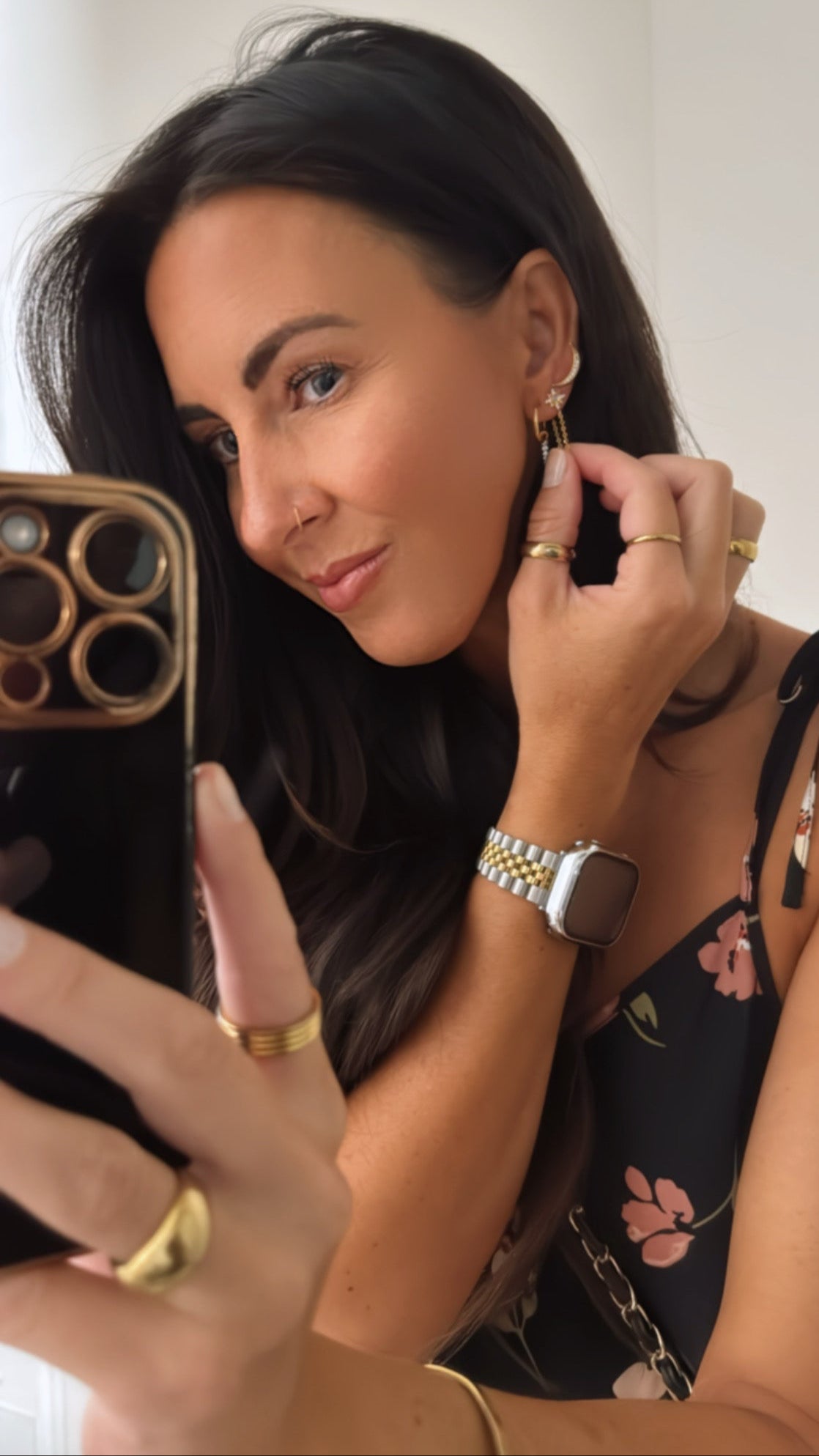 Woman adjusting earrings in front of a phone camera, wearing multiple rings and a watch.